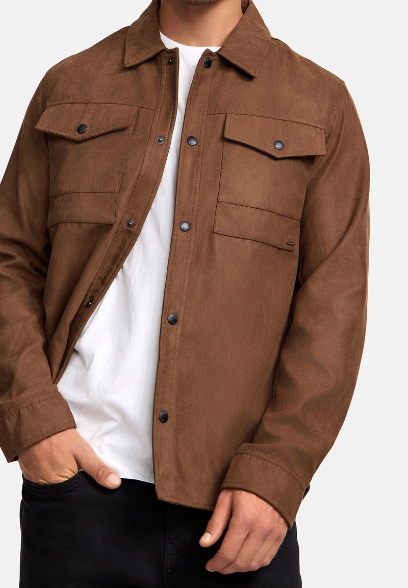 Homme portant une veste à boutons marron avec des poches à rabat sur un t-shirt blanc et un pantalon noir, la main dans la poche, contre un fond uni.