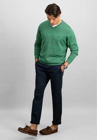 Grøn v-hals sweater, marineblå chinos og brune loafers. Sweateren har et diskret logo; chinosene har lige snit og opkrøllede manchetter.