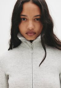 Svijetlo siva zip-up krvavi s visokom ovratnikom i rebrastom teksturom. Ima srebrni patentni zatvarač i glatku tkaninu. Jednostavan, minimalistički dizajn.
