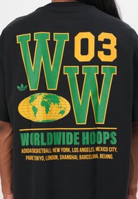 Svart t-skjorte med store grønne og gule grafikker: "WOW 03", en globe, og teksten "WORLDWIDE HOOPS" med bynavn i hvitt.
