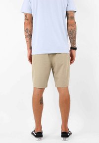 Shorts beiges en tissu léger, avec une coupe droite et une longueur jusqu'aux genoux. Associés à un t-shirt bleu clair et des baskets noires.