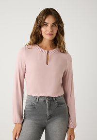 Langärmlige pinke Bluse mit einem gefältelten Rundhalsausschnitt und einem vorderen Schlitz, kombiniert mit hoch geschnittenen grauen Jeans. Glatte Textur und taillierte Passform.