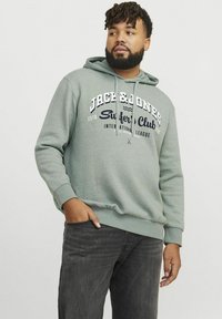 Sudadera verde con bolsillo tipo canguro, que presenta una impresión de "Jack & Jones Surfer's Club" en blanco y azul marino. Combinada con jeans oscuros.