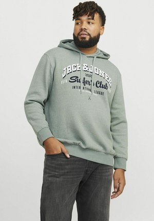 Zelena dukserica s kenguruvim džepom, s natpisom "Jack & Jones Surfer's Club" u bijeloj i mornarskoj plavoj boji. Uparena s tamnim trapericama.