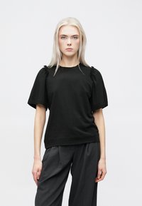 Fekete, rövid ujjú blúz, vállain ráncolt díszítéssel, puha anyagból készült. Szürke, bő szárú nadrággal párosítva, fehér háttér előtt bemutatva.