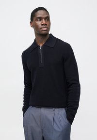 Tmavomodrý pletený pulóver s plochým golierom a polovičným zipsom na výstrihu. Texturovaná látka a priliehavé rukávy, v kombinácii s svetlo šedými plisovanými nohavicami.