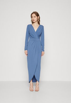 Robe longue bleue en tissu lisse avec un décolleté en V, manches longues, plis à la taille et une fente sur le côté, associée à des talons argentés à lanières.