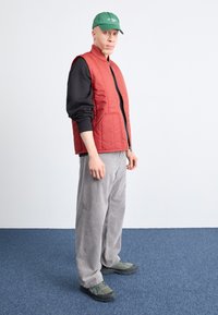 Colete acolchoado vermelho sobre uma camisa preta de mangas longas, calças de veludo cotelê cinzas e um boné verde. O calçado são ténis atléticos cinza e preto.