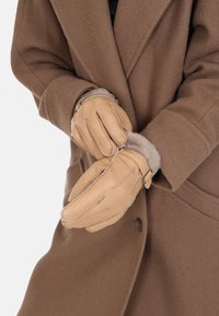 Tan leather gloves with fur lining, featuring a cinched wrist and decorative stitching, worn over a brown coat.