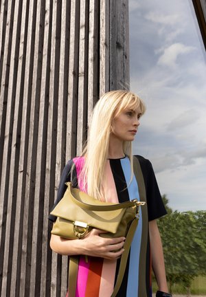 Frau mit langem blondem Haar hält eine olivgrüne Handtasche, trägt ein bunt gestreiftes Kleid und steht neben einer Holzwand und einem Glasfenster.