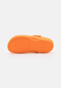 Suela de zapato de goma naranja con un destacado patrón de banda de rodadura, logo circular y detalles de correa ajustable a lo largo del costado. Superficie plana y texturizada.