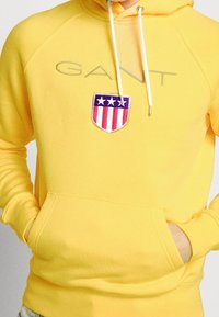 Žlutá mikina s kapucí na stažení, přední kapsou a výrazným vyšívaným štítovým logem s červenými a modrými pruhy a bílými hvězdami.