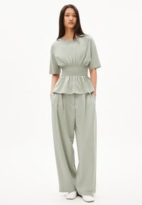 Hellgrünes, lässig geschnittenes Oberteil mit gerafftem Bund und kurzen Ärmeln, kombiniert mit weiten Hosen. Glatte Textur, minimales Design und keine Muster.