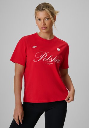 Kobieta o blond włosach, ubrana w czerwoną koszulkę Polska i czarne spodnie, stojąca na jednolitym szarym tle.