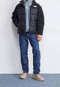 Persona vistiendo una chaqueta acolchada negra, sudadera gris de Nike, jeans de mezclilla azules con bolsillos de parche y zapatillas beige, de pie sobre una alfombra azul.