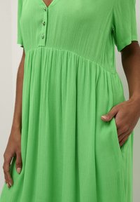 Grünes, kurzärmeliges Kleid aus strukturiertem Stoff. Es verfügt über einen V-Ausschnitt und drei Knöpfe vorne sowie einen ausgestellten Rock und Seitentaschen.