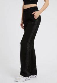 Black sweatpants with a drawstring waist, featuring side pockets and a tonal logo pattern along the outer leg. Soft, stretchy fabric.
