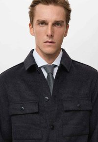 Dunkle Jacke aus Wollmischung mit Kragen, zwei Vordertaschen und Knopfverschluss. Trägt über einem hellblauen Hemd und einer grauen Krawatte.