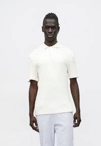 Polo de manga curta de cor creme com textura canelada e colarinho clássico. O tecido parece macio e ligeiramente elástico.