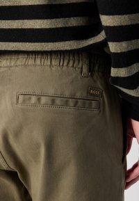 Pantaloni kaki cu talie elastică, având un buzunar pe spate și o etichetă de marcă. Materialul are o suprafață netedă, ușor texturată.