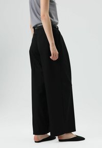 Zwarte, wijde broek met hoge taille, gladde stof, zijslits en lussen voor een riem. Gecombineerd met een grijs topje en zwarte instapschoenen.