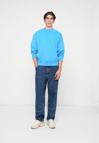 Sudadera azul claro con puños y dobladillo acanalados, combinada con jeans oscuros de corte holgado que presentan costuras curvas. Zapatillas blancas completan el look.