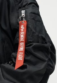 Μανίκι μαύρου μπουφάν με τσέπη με φερμουάρ και προσαρτημένη κόκκινη υφασμάτινη ετικέτα που γράφει «ΑΦΑΙΡΕΣΤΕ ΠΡΙΝ ΤΗΝ ΠΤΗΣΗ», κρεμασμένη από το τράβηγμα του φερμουάρ.