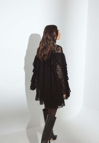 Juoda nėriniai suknelė su sluoksniuotais volanais ir ilgomis rankovėmis, derinama su juodomis aukštakulnėmis batų iki kelių, matoma iš nugaros prieš baltą foną.