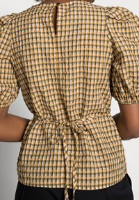 Blusa a quadri giallo senape e bianco, con maniche a palloncino, vita con lacci e chiusura a oblò sul retro. Tessuto di cotone testurizzato.
