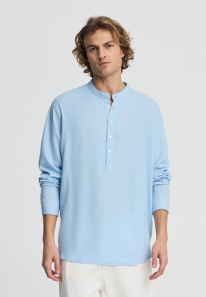 Giovane con capelli ricci che indossa una camicia azzurra a maniche lunghe abbottonata e pantaloni bianchi, in piedi contro uno sfondo semplice.