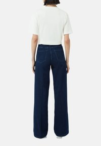 Weißes T-Shirt mit kurzen Ärmeln, kombiniert mit dunkelblauen weit geschnittenen Jeans, die über zwei Gesäßtaschen und sichtbare Nähte verfügen.