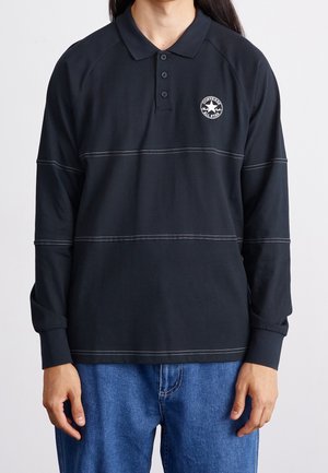 Osoba nosząca czarną koszulkę polo z długim rękawem z białym przeszyciem i logo Converse All Star, w połączeniu z niebieskimi dżinsami.