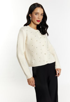 Weißer Strickcardigan mit Perlenbesatz, rundem Ausschnitt und langen Ärmeln. Kombiniert mit schwarzen Hosen. Einfaches und strukturiertes Design.