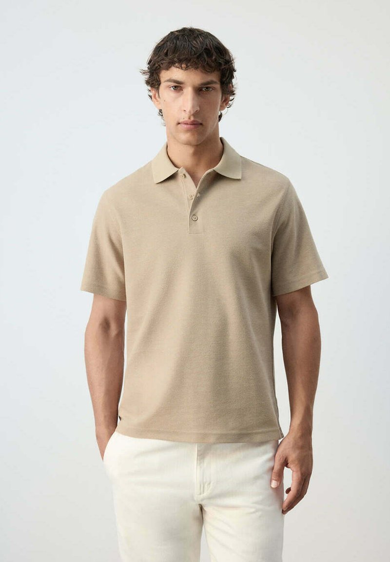 Jeune homme aux cheveux bouclés foncés portant un polo beige à manches courtes et un pantalon blanc sur un fond clair uni.