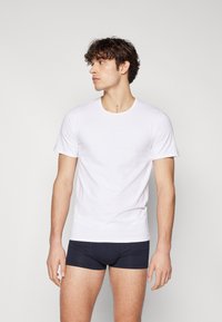 Bílý krátký rukáv tričko, kulatý výstřih, hladká látka. Nosené s tmavě modrým úzkým spodním prádlem. Model stojí v mírném úhlu.