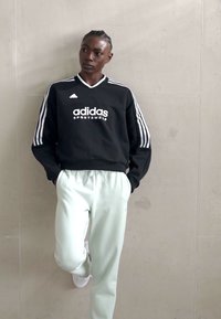 Sweatshirt preto da Adidas com riscas brancas, logótipo no peito; calças de treino verde claro, detalhadas com cintura elástica; sapatos brancos.