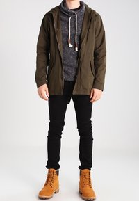 Olivgrüne Kapuzenjacke über einem grauen Strickpullover, kombiniert mit schwarzen Skinny Jeans und hellbraunen Schnürboots. Verfügt über Kordelzug und Taschen.