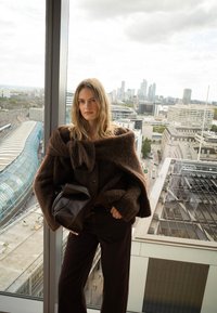 Vrouw met lang blond haar, gekleed in een oversized bruine trui en donkere broek, met een zwarte tas in de hand, staand bij een groot raam met uitzicht op de stad.