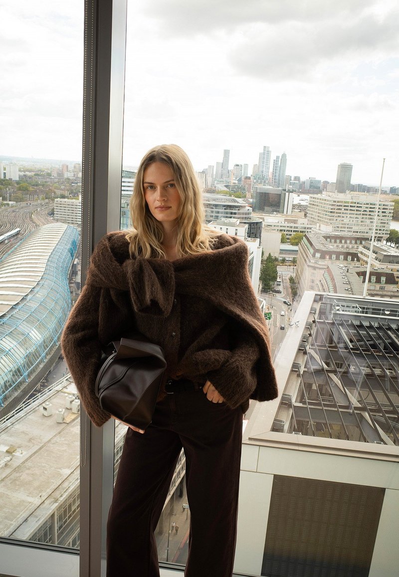 Vrouw met lang blond haar, gekleed in een oversized bruine trui en donkere broek, met een zwarte tas in de hand, staand bij een groot raam met uitzicht op de stad.