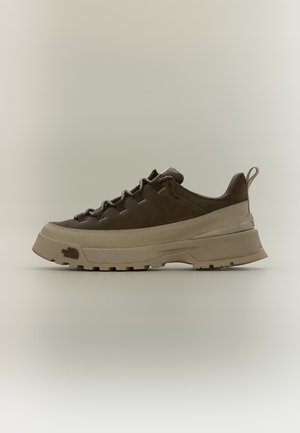 Brauner niedriger Wanderschuh mit dicker beiger Sohle, Schnürdesign und Zuglasche an der Ferse vor beigem Hintergrund.