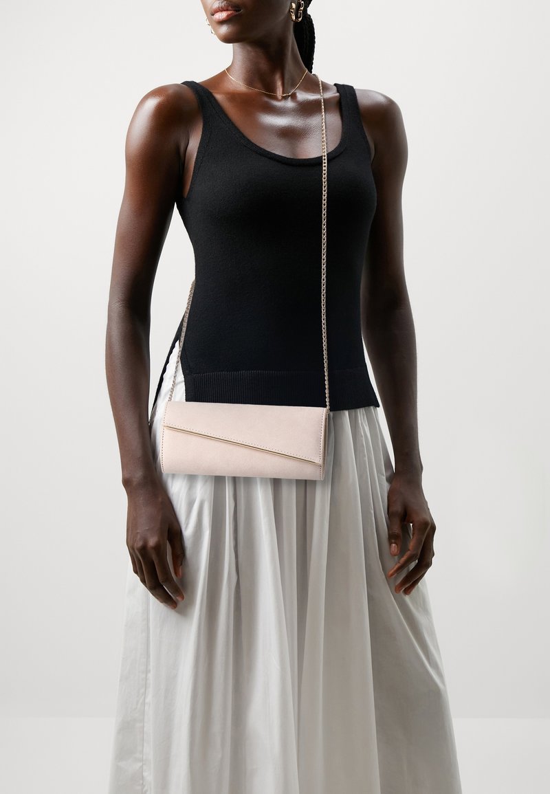 Femme à la peau foncée portant un haut noir sans manches et une jupe plissée blanche, tenant une pochette enveloppe rose clair avec une bandoulière chaîne.