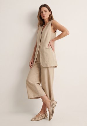 Frau steht mit einer Hand in der Hüfte, trägt beige ärmellosen Blazer, passende weite, kurze Hosen und geflochtene Mary Jane Ballerinas.