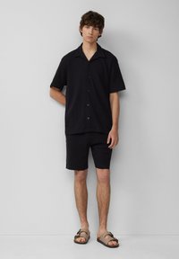 Chemise à manches courtes, boutonnée, et shorts assortis en tissu noir texturé, associés à des sandales marron clair avec une sangle décorative.