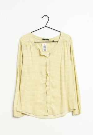 Blouse jaune à manches longues avec un motif à carreaux subtil, suspendue sur un cintre noir sur un fond blanc, avec une étiquette visible.