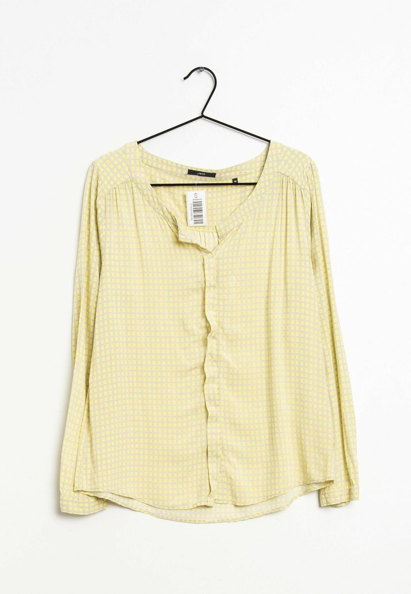 Blouse jaune à manches longues avec un motif à carreaux subtil, suspendue sur un cintre noir sur un fond blanc, avec une étiquette visible.