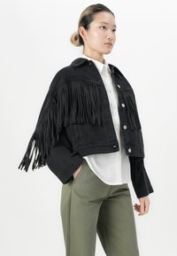 Giovane donna che indossa un giubbotto di jeans nero con frange sopra una camicia bianca e pantaloni verde oliva, in piedi davanti a uno sfondo bianco uniforme.