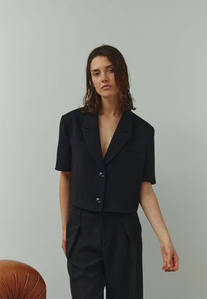 Fekete, rövid ujjú, rövidített blézer két első zsebbel és fényes gombokkal. A modellen hozzáillő fekete nadrág látható, semleges háttér előtt.