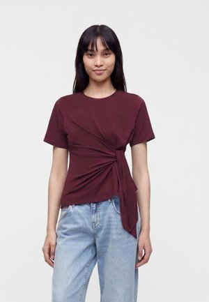 Jeune femme aux longs cheveux foncés, portant un haut bordeaux à manches courtes avec un détail noué sur le côté et un jean en denim bleu clair.