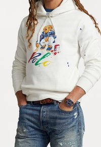Vit hoodie med en björngrafik i färgglada färgsprut, tillsammans med slitna blå jeans och ett brunt läderarmband.