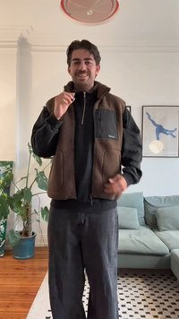 Brauner Fleece-Weste mit einer schwarzen Reißverschlusstasche, getragen über einem schwarzen Langarmshirt. Lockere schwarze Hose. Inneneinstellung mit Grünpflanzen.